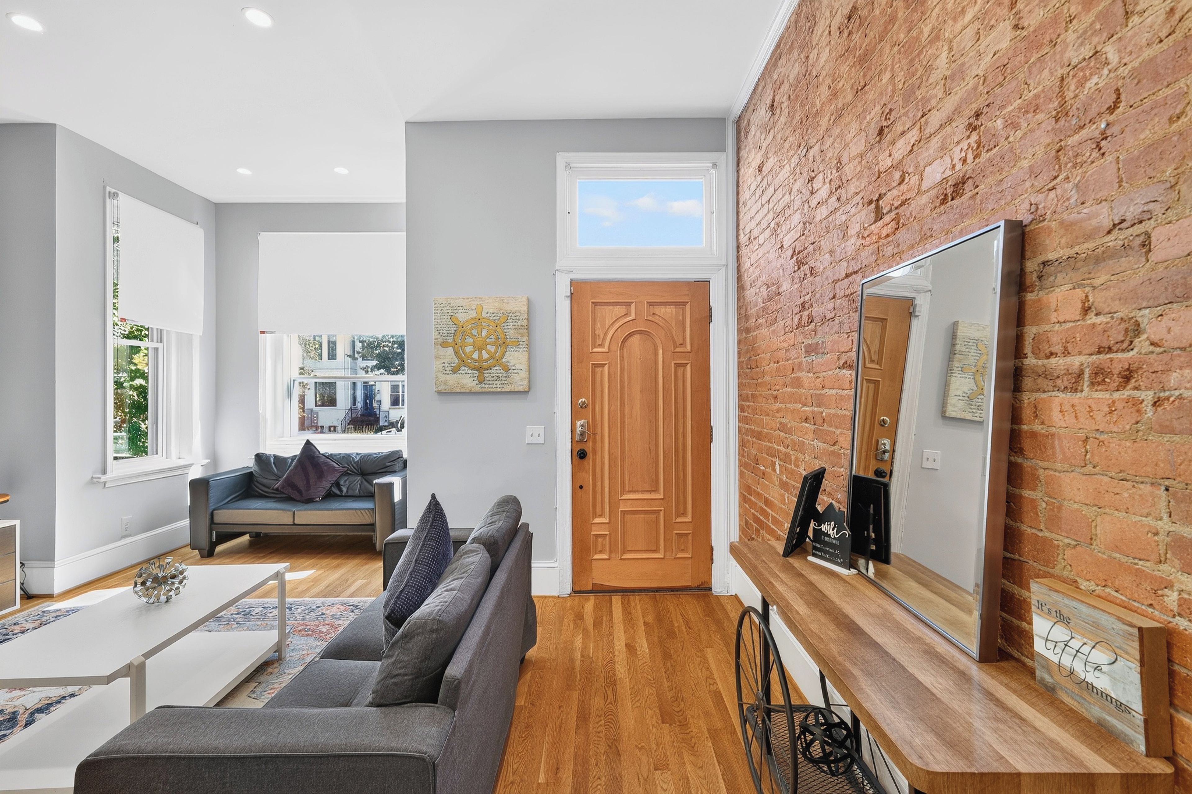 A modern living room with a grey sofa, wooden flooring, a brick accent wall, and a large mirror.