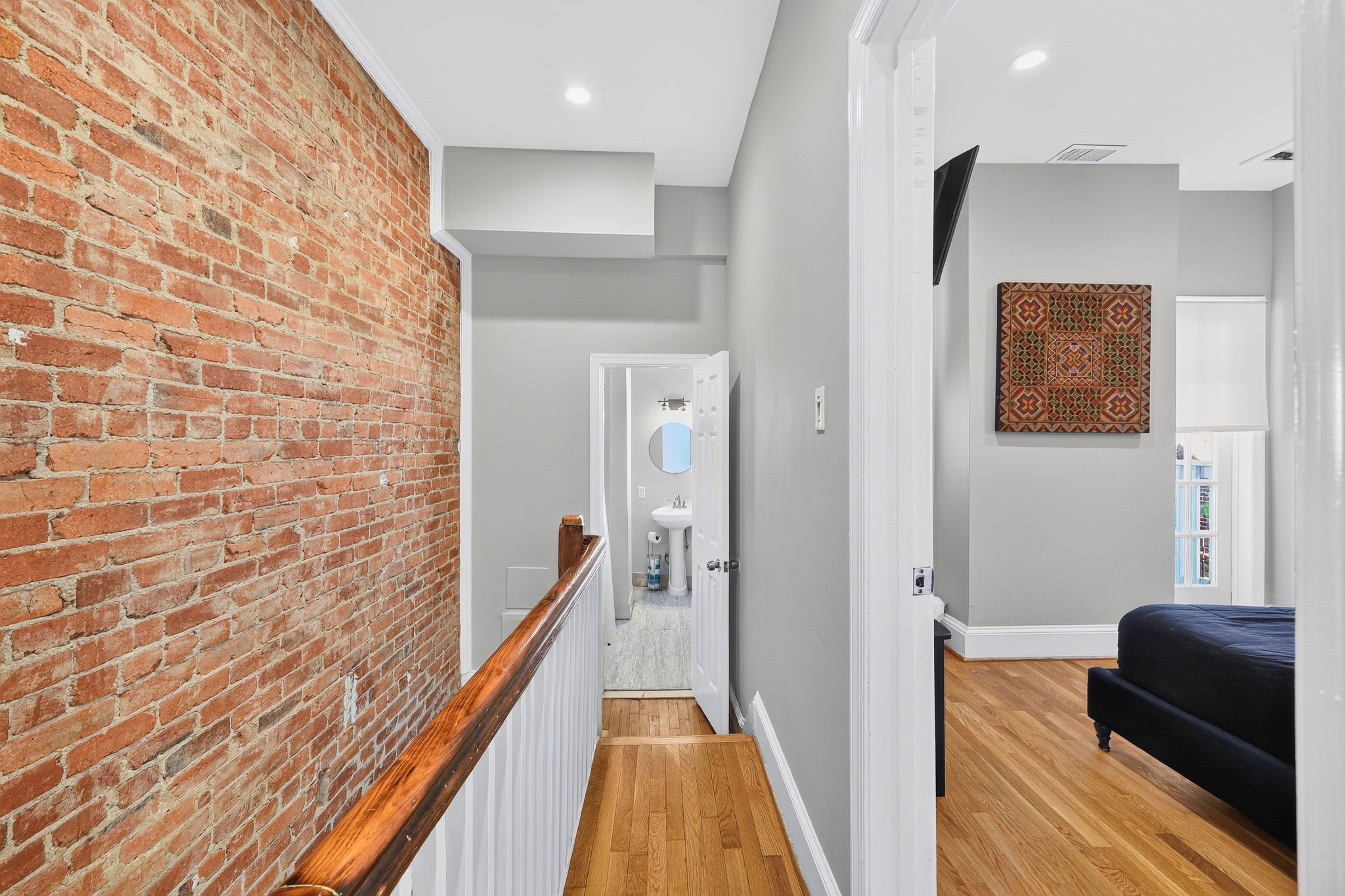 A hallway with a brick wall, wooden floor, and a bed in the adjacent room.