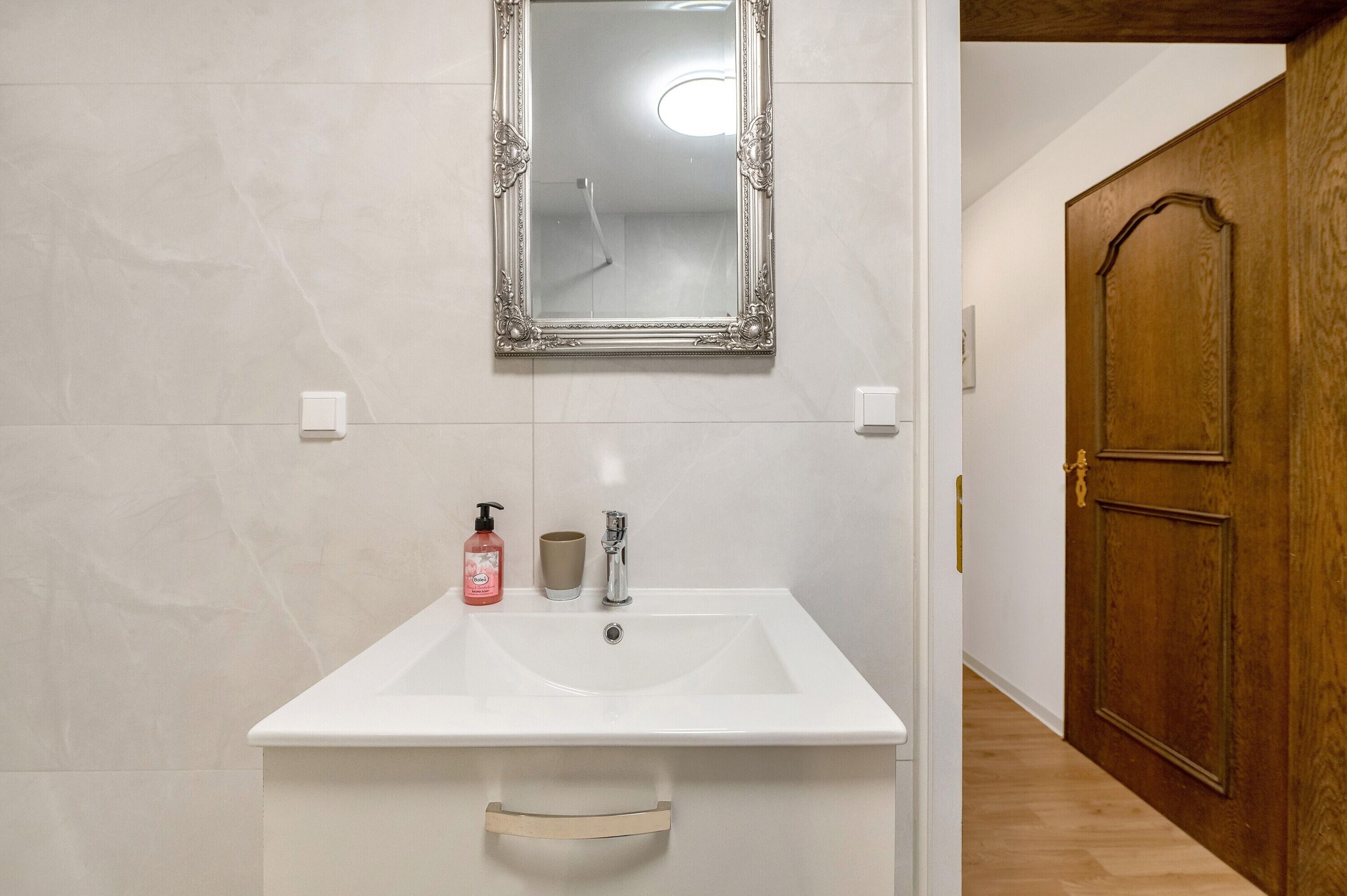 Une salle de bain avec un lavabo blanc, un miroir, un flacon de savon et une porte en bois.