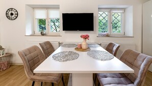 A dining room with a white table, six chairs, a flat-screen TV, and windows with a view of greenery. - Apartment 'Green House' with Private Terrace, Private Garden and Wi-Fi (Neunkirchen)