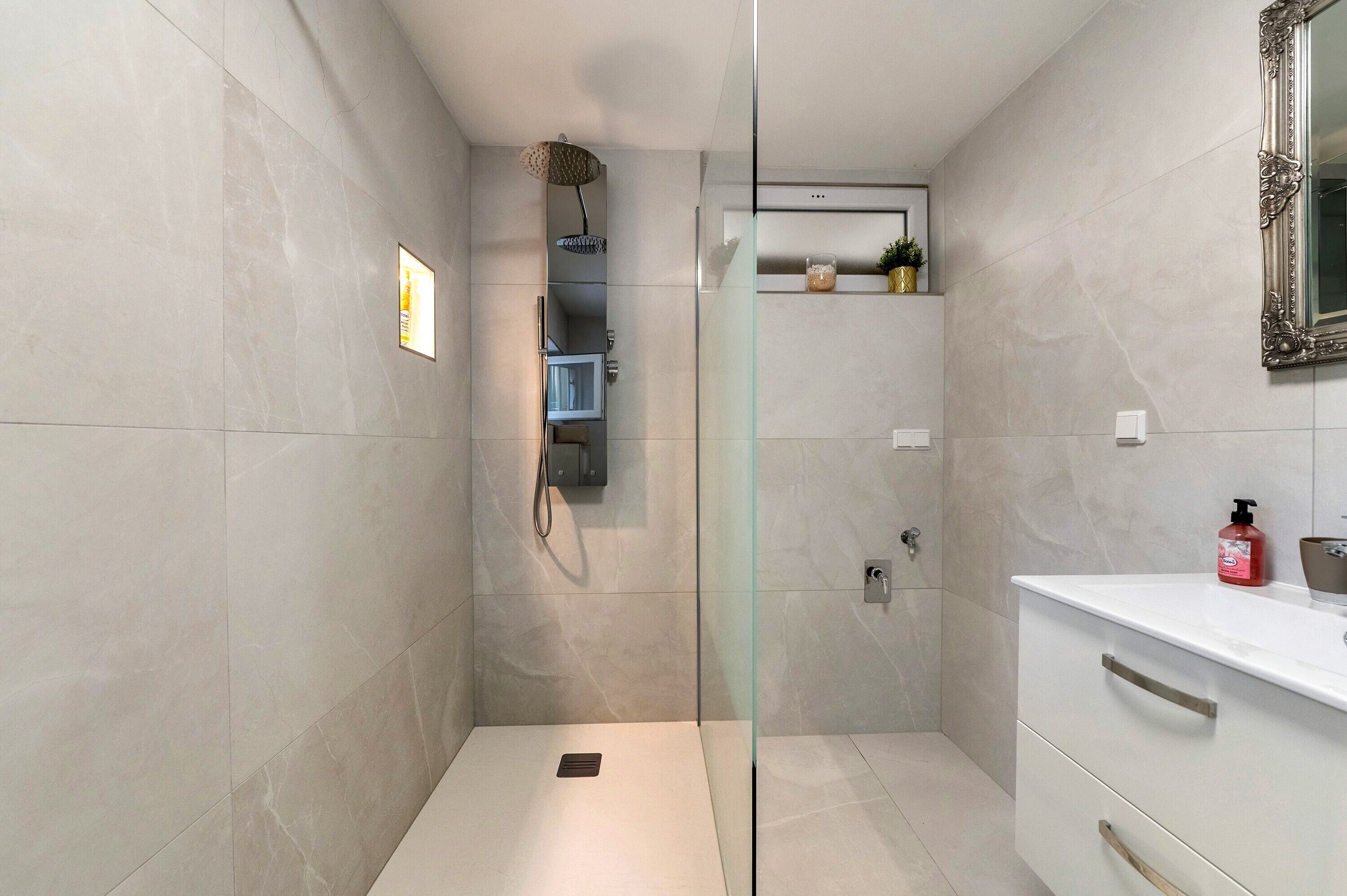 A modern bathroom with a glass-enclosed shower, a white vanity with a mirror, and a small shelf with toiletries.