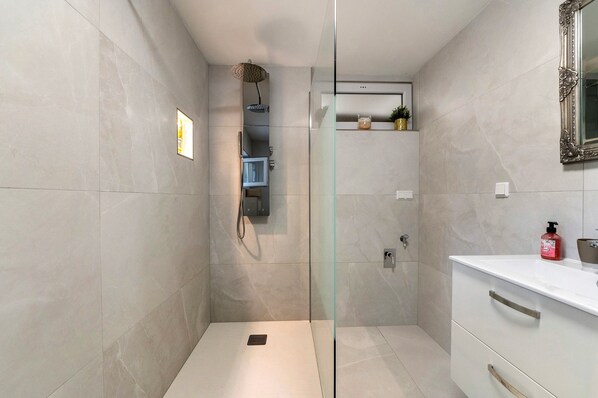 A modern bathroom with a glass-enclosed shower, a white vanity with a mirror, and a small shelf with toiletries.