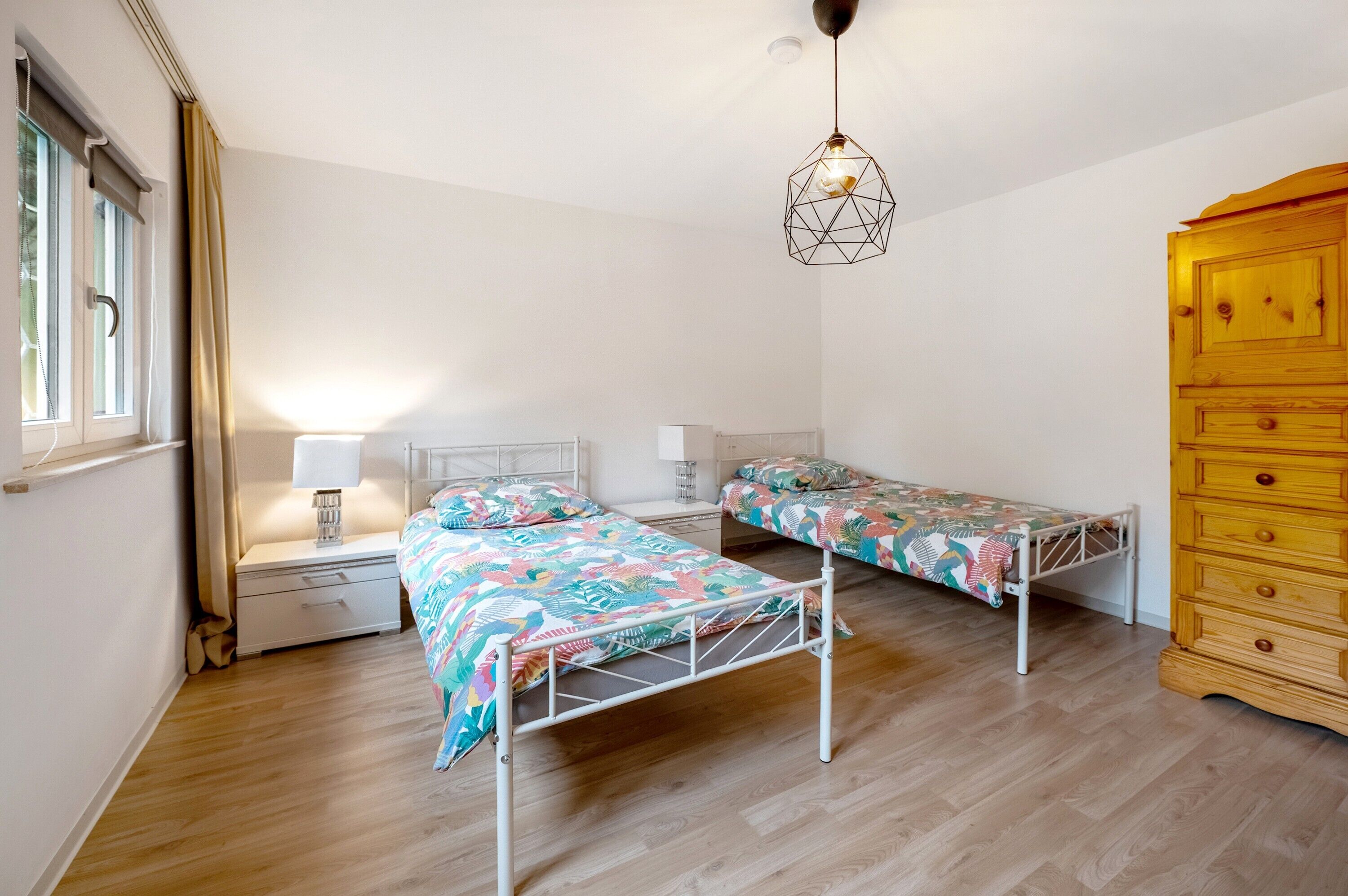Une chambre avec deux lits simples, une commode en bois, une table de chevet avec une lampe et une fenêtre avec des rideaux.
