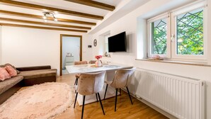 A modern living room with a dining area, wooden beams on the ceiling, a white radiator, and a window with a view of greenery. - Apartment 'Green House' with Private Terrace, Private Garden and Wi-Fi (Neunkirchen)
