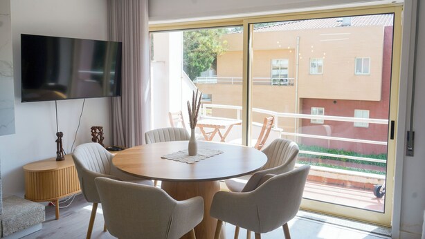Un salon moderne avec un téléviseur à écran plat, une table ronde en bois entourée de chaises, un vase et une vue sur un immeuble par une porte vitrée.