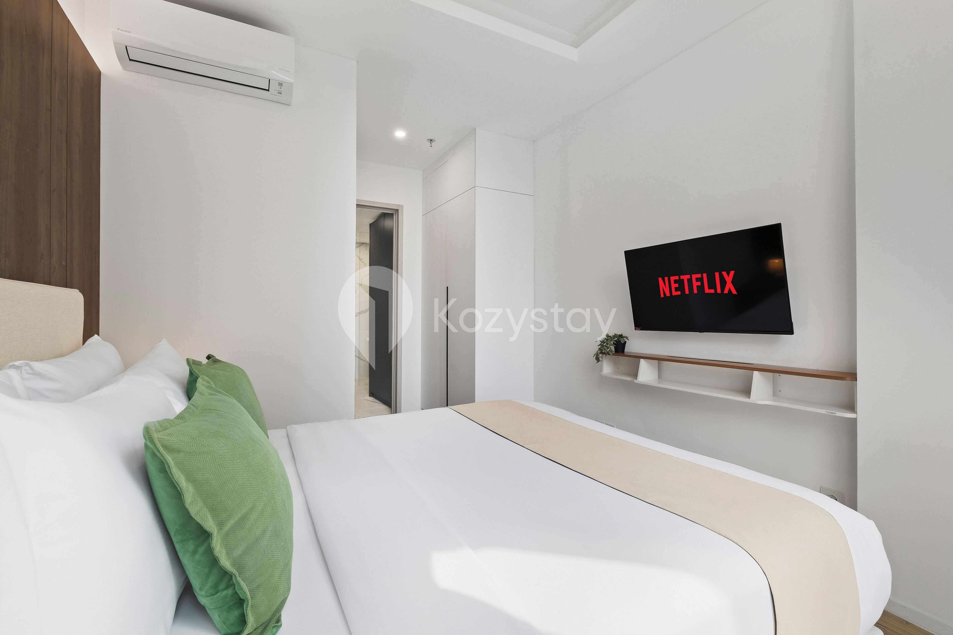 A modern hotel room with a bed, a mounted TV displaying 'NETFLIX', and a small shelf with a plant.
