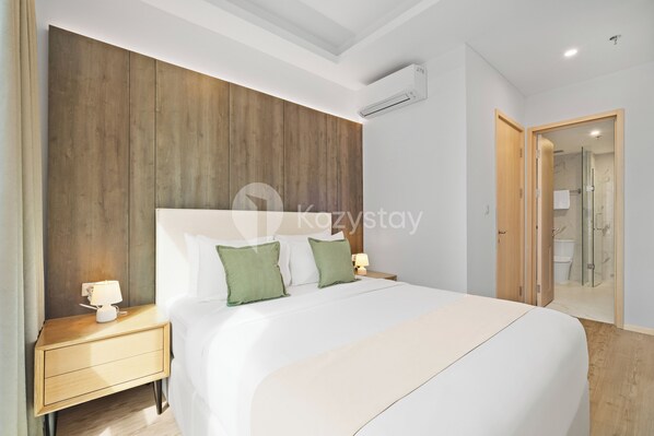Une chambre d’hôtel moderne avec une tête de lit en bois, un grand lit avec une literie blanche, deux tables de chevet avec des lampes et une porte de douche en verre.