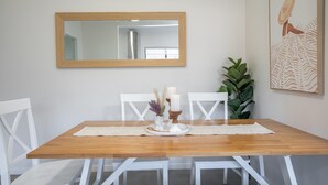 A dining area with a wooden table, white chairs, a mirror, a plant, and a framed artwork.