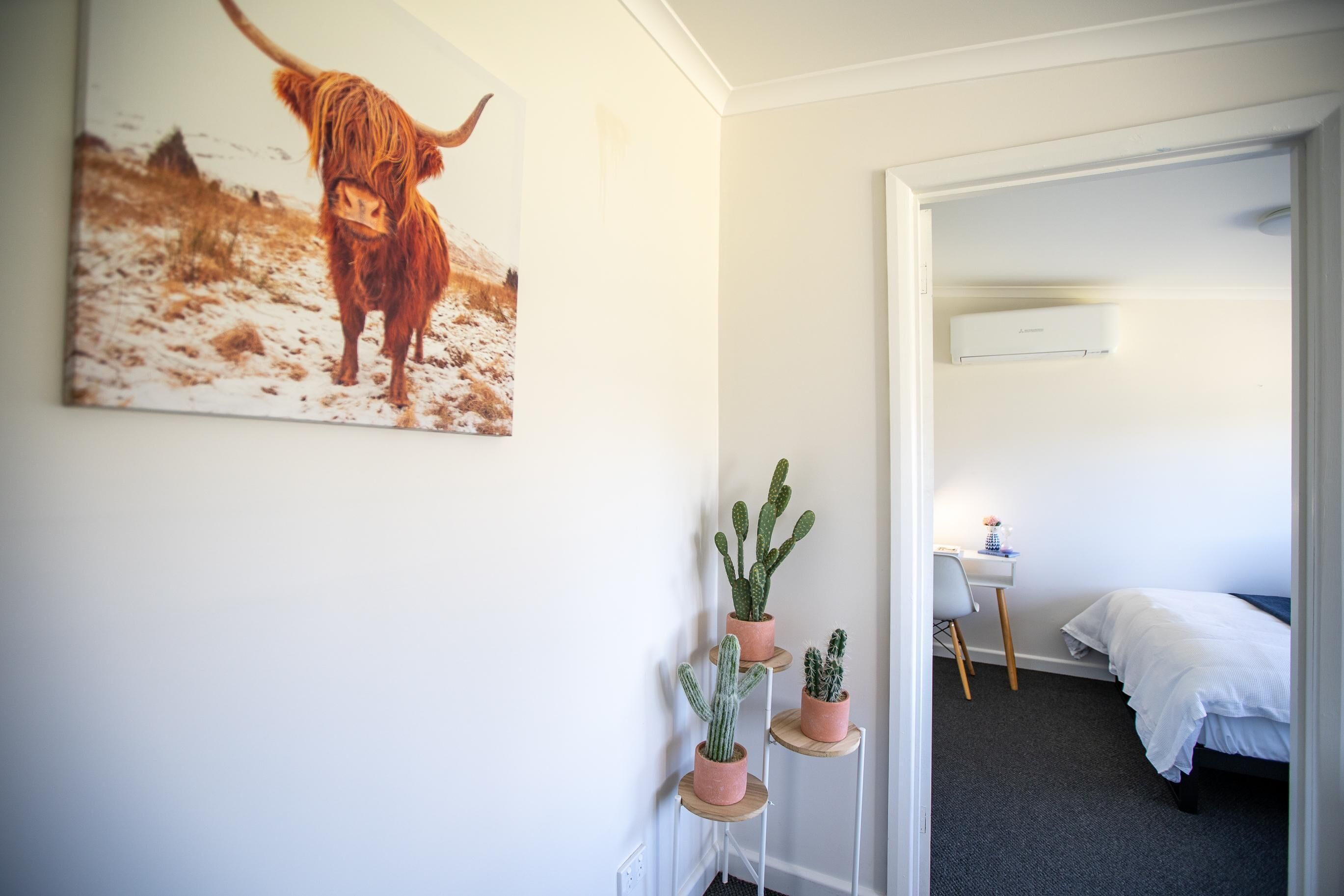 A hotel room with a bed, a desk, and a chair. There is a painting of a cow on the wall and three potted plants on a shelf.