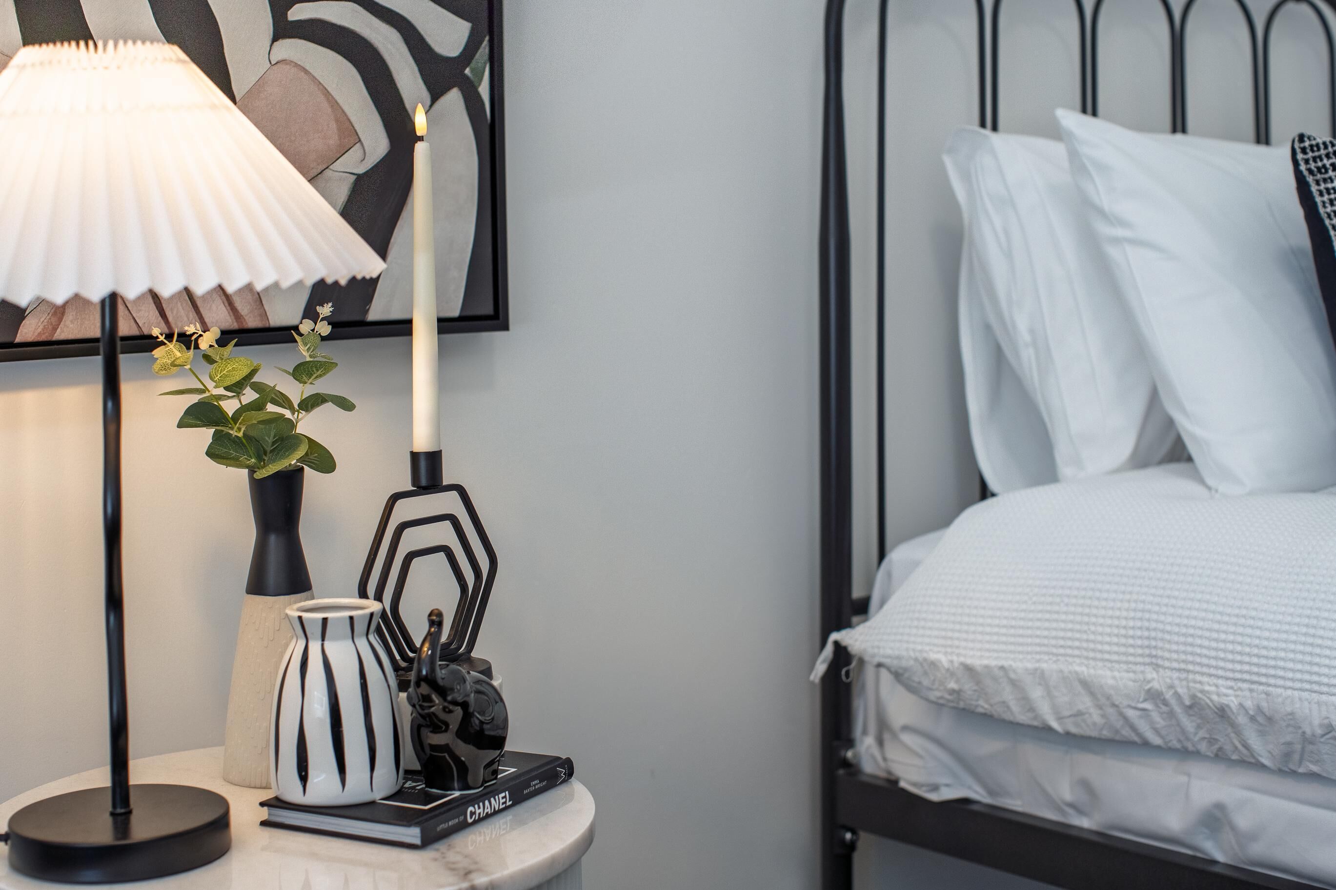 A neatly arranged bedroom with a bed, a bedside table with a lamp, a vase with flowers, and a framed picture on the wall.