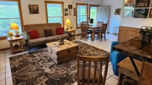A cozy living room with a sofa, coffee table, and dining area. There are two windows with blinds, a ceiling fan, and a wall with framed pictures.