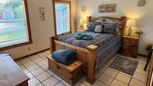 A bedroom with a wooden bed, a bench, a nightstand, a lamp, and a window with blinds.