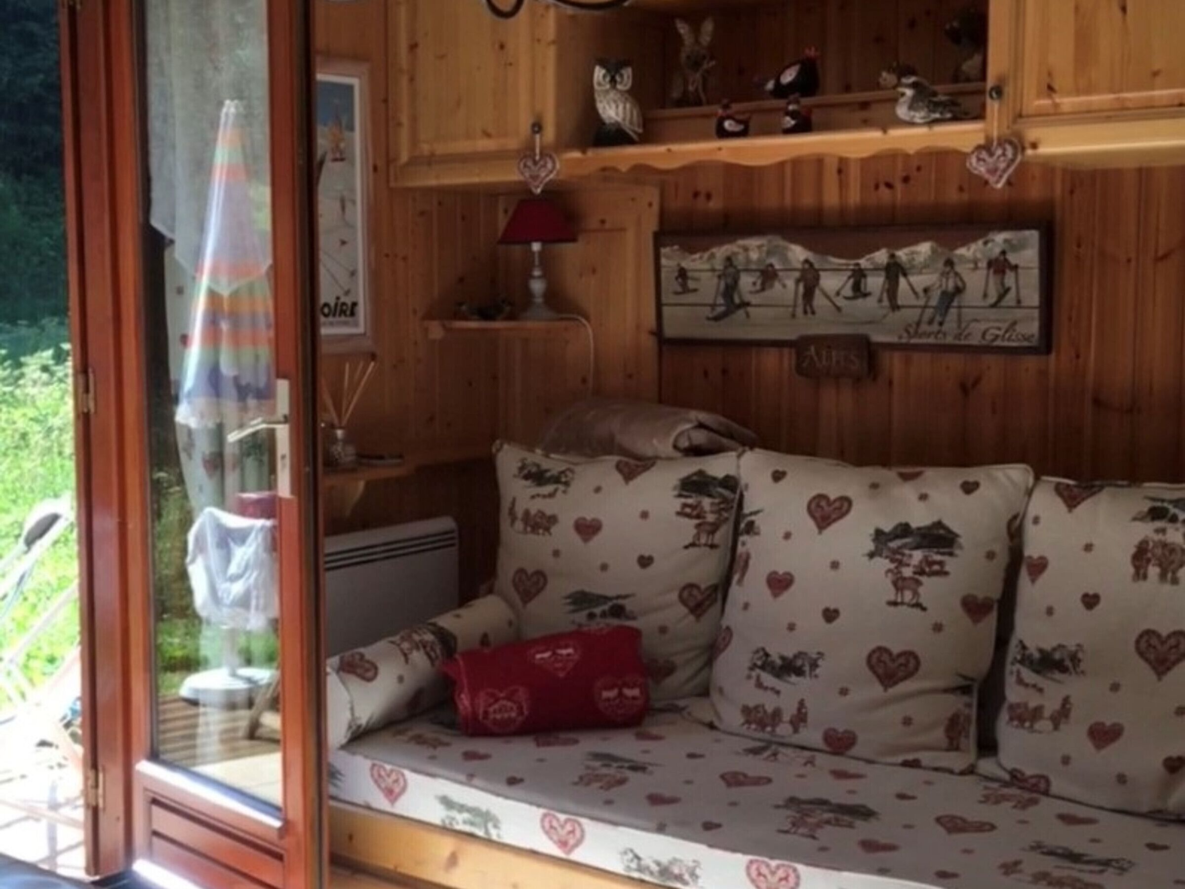 Une pièce chaleureuse avec un décor intérieur en bois, un canapé agrémenté de coussins à motifs en forme de cœur, une petite table avec une lampe et une étagère ornée d’objets décoratifs.