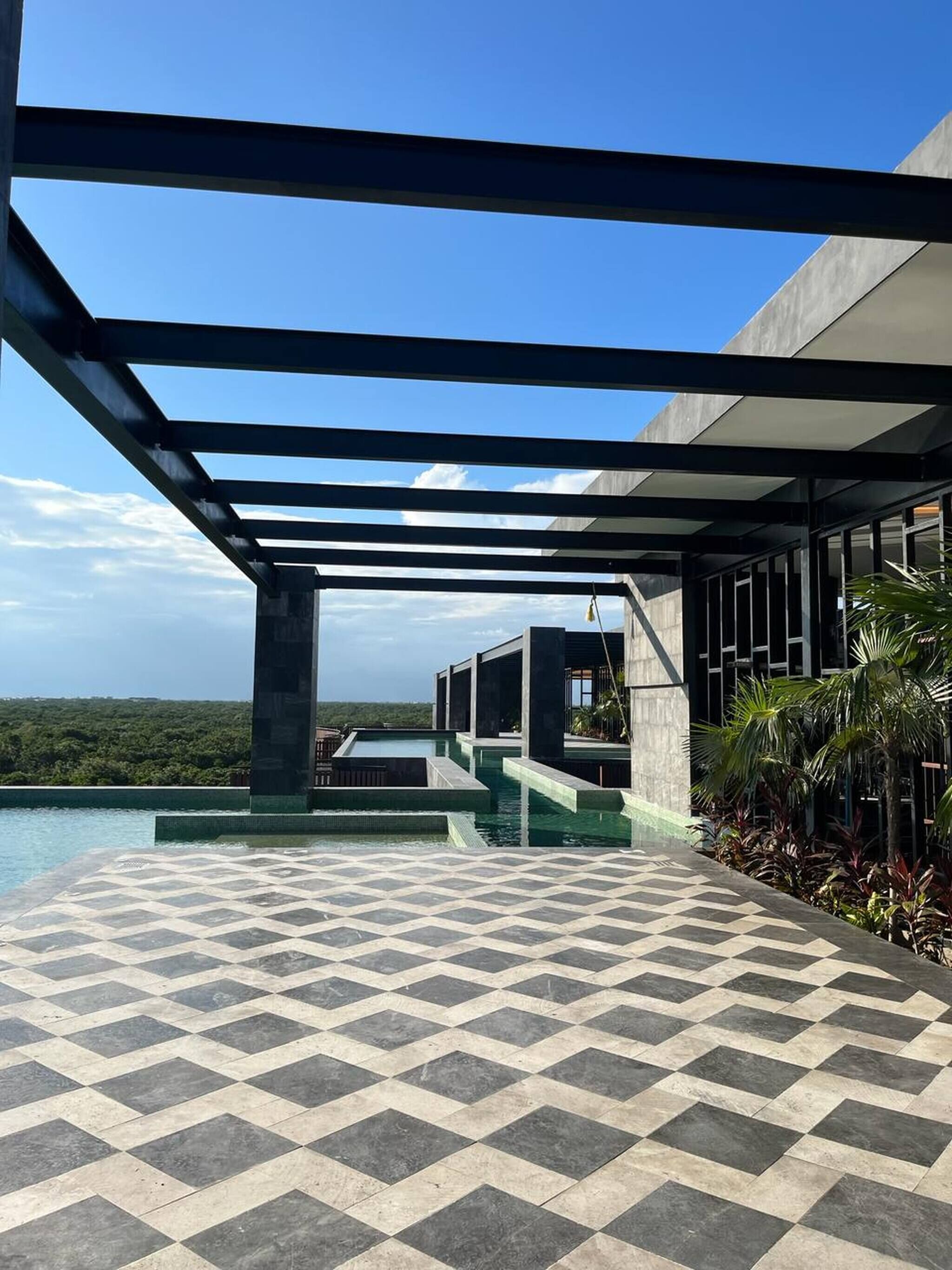 A modern outdoor pool area with a checkered floor, a covered seating area, and a view of a lush landscape.