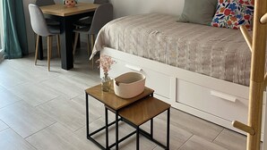Un dormitori modern amb un llit, un escriptori i una cadira. Hi ha un rellotge a la paret i una taula petita amb un vas i una cistella.