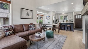 Ein großzügiges Wohnzimmer mit einer braunen Ledersofa, einem runden Couchtisch und einem grünen Hocker. Im Hintergrund ist ein Essbereich mit Holztisch und Stühlen zu sehen.