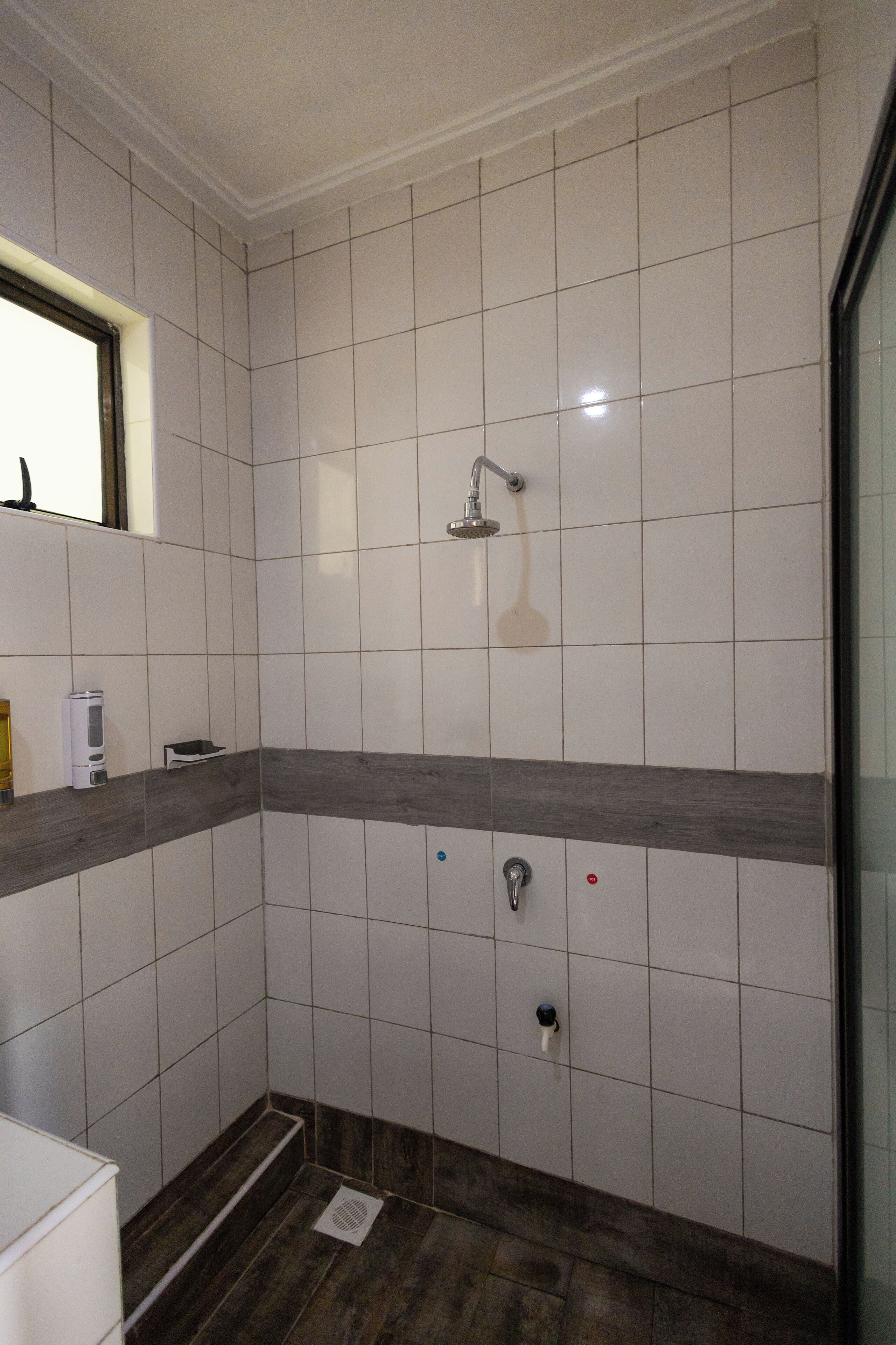 Une salle de bain carrelée de blanc, avec une douche, un plancher en bois et une petite étagère où sont rangés les articles de toilette.