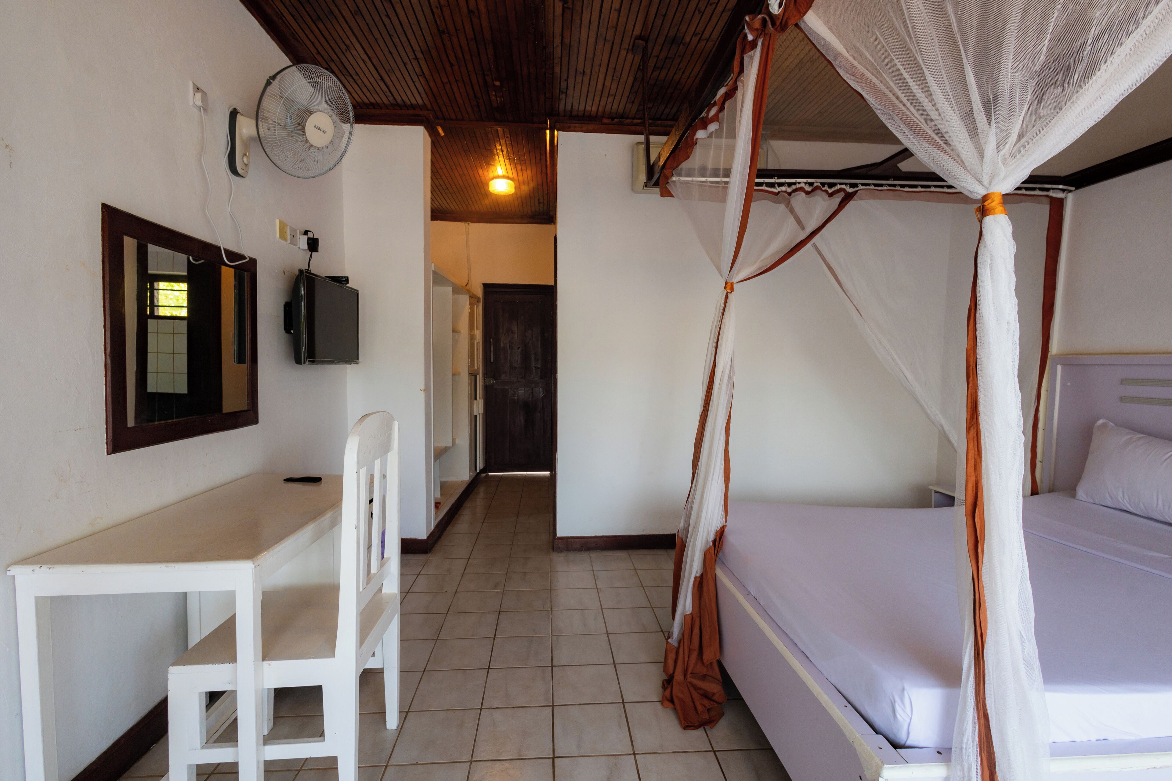 Quarto com cama, escrivaninha, cadeira, televisão e ventoinha no teto.