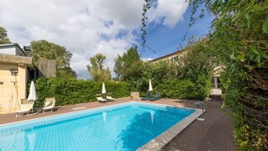 A rectangular swimming pool with a tiled edge, surrounded by a brick patio. There are lounge chairs and umbrellas along the poolside. - Achillea Apartment with Shared Pool and Garden (Rosignano Marittimo)