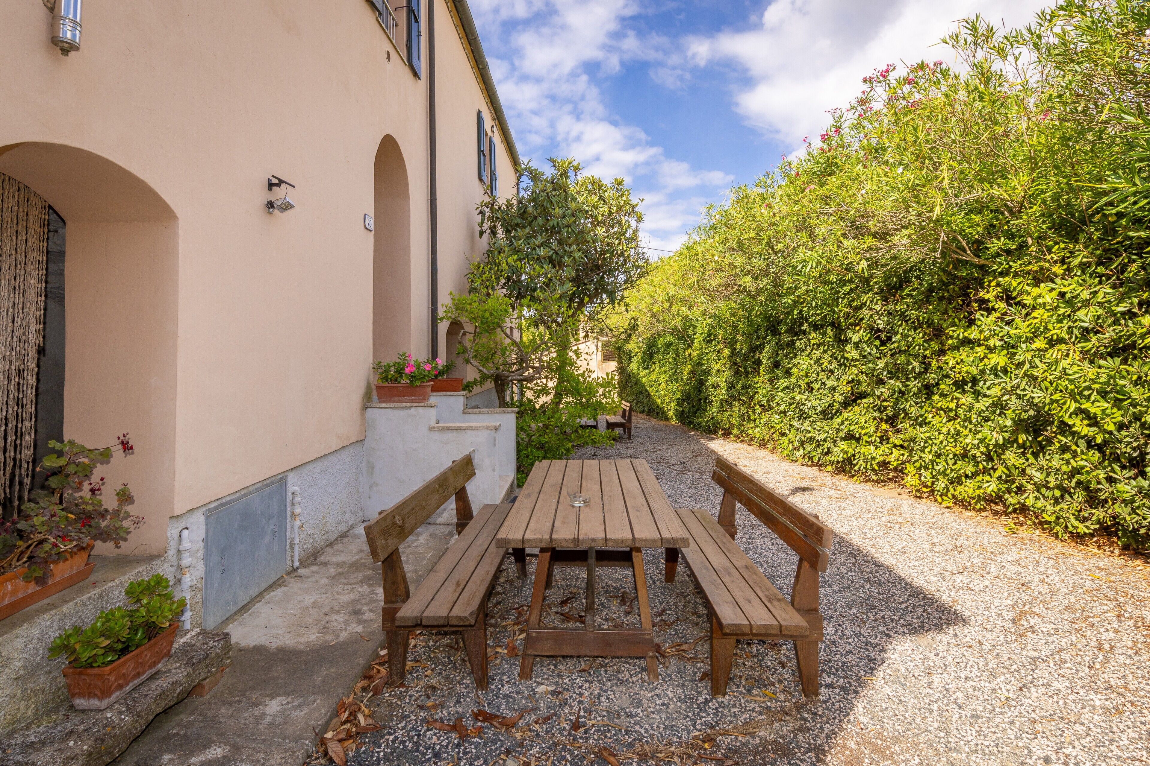 Une terrasse avec une table en bois et des bancs, entourée d’un chemin de gravier et de verdure.