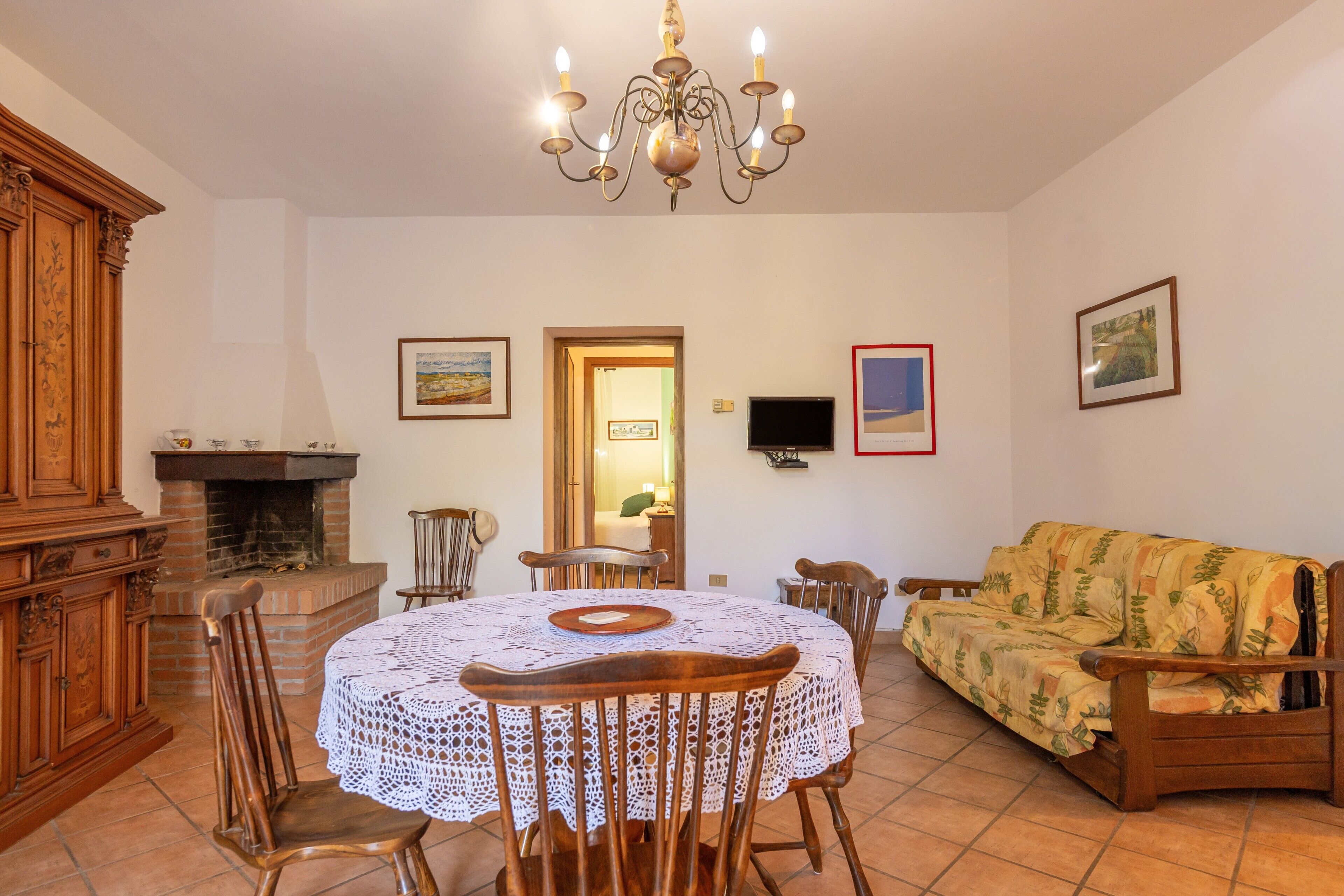 A dining area with a round table, chairs, a chandelier, a fireplace, a sofa, and framed pictures on the walls.