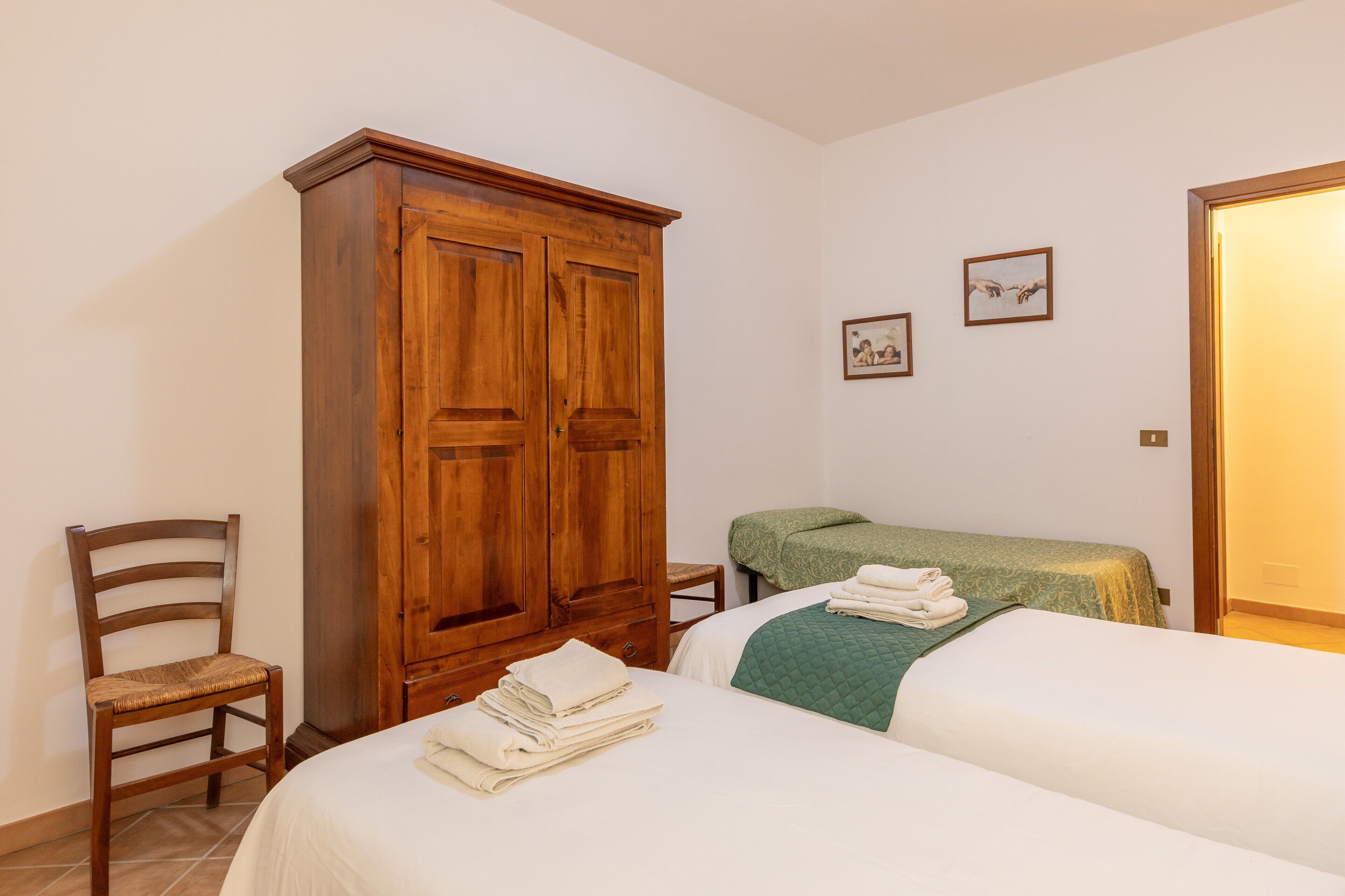 Een slaapkamer met een houten garderobe, een enkel bed met een groene deken, een stoel en twee ingekaderde foto's aan de muur.