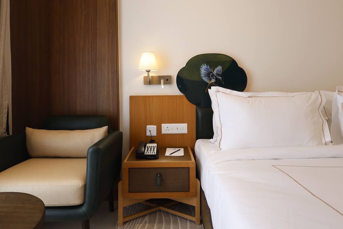 A hotel room with a bed, a chair, a small table with a lamp, and a nightstand with a phone and a decorative item.