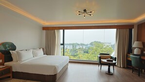 Ein Hotelzimmer mit einem großen Bett, einem Schreibtisch, einem Sessel und Blick auf die Stadt durch große Fenster.