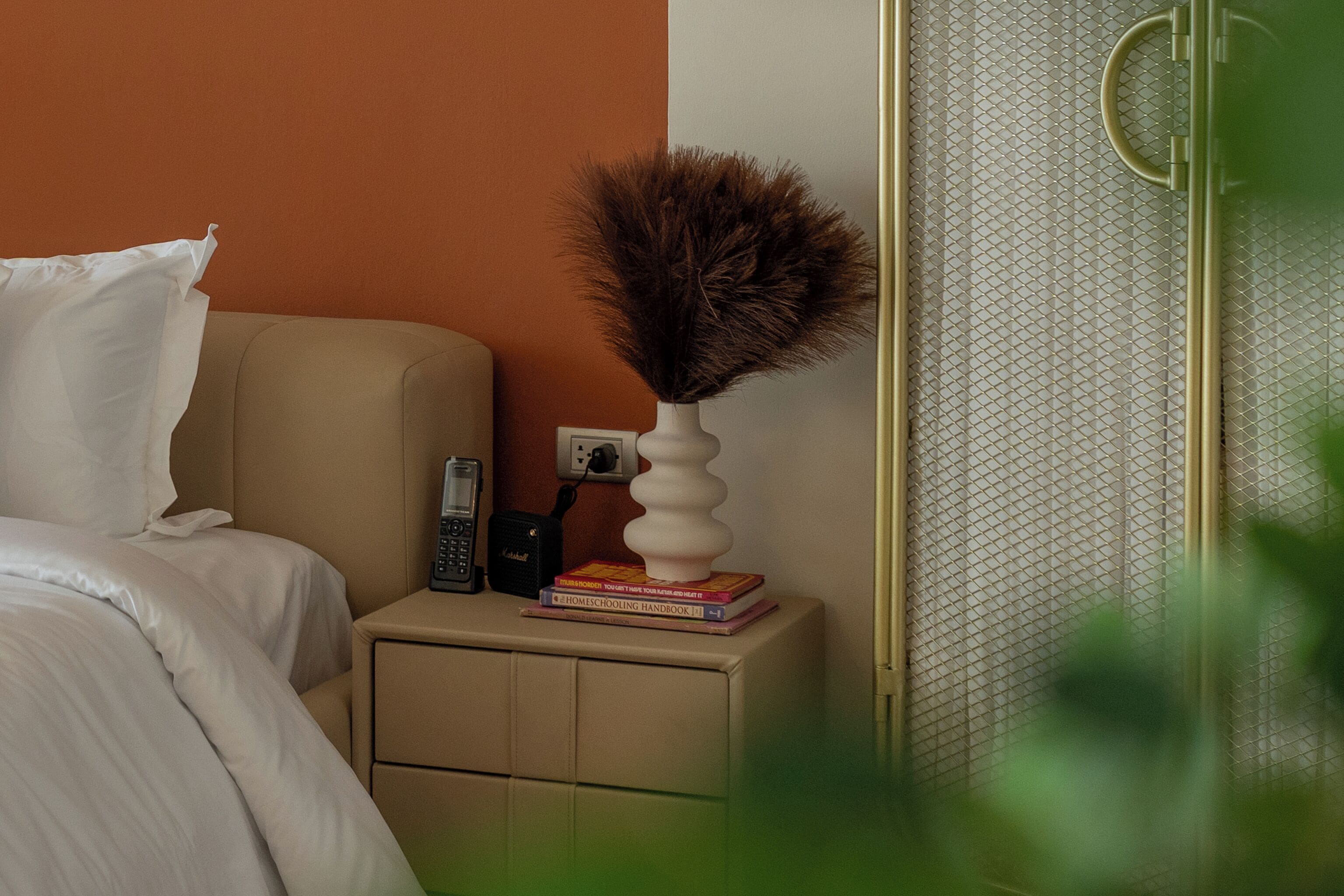 A neatly made bed with white linens, a beige nightstand with a white vase and books, and a telephone.