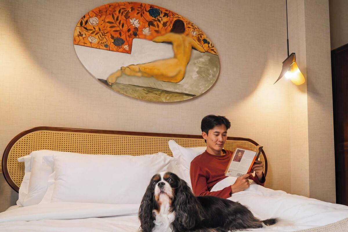 A hotel room with a bed, a reading lamp, a painting on the wall, and a dog.