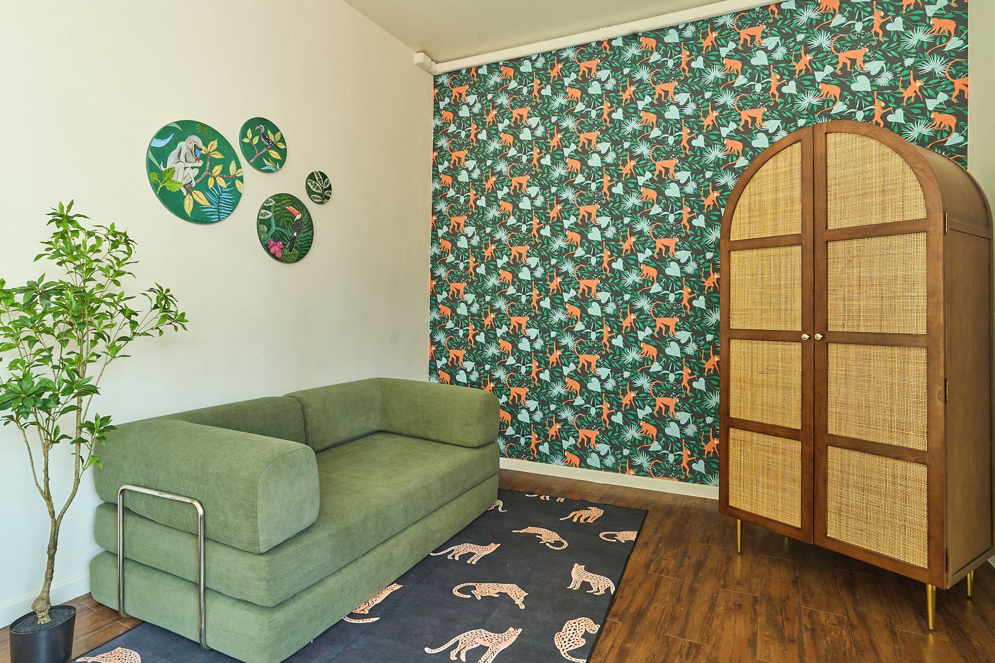 Una habitación con un sofá verde, un mueble con puertas de mimbre, una pared con papel tapiz de temática selvática y una planta pequeña.