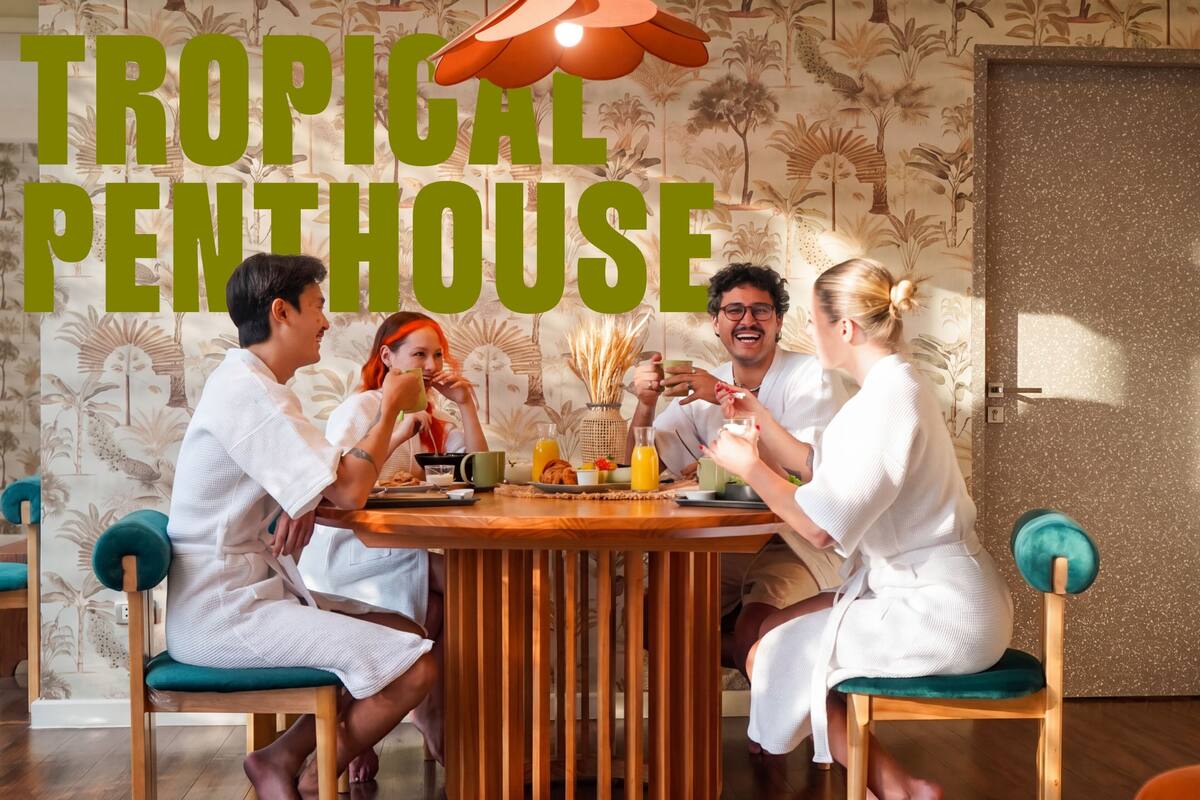 A group of people seated around a wooden table in a room with a tropical-themed wallpaper and a large sign reading 'TROPICAL PENTHOUSE'.