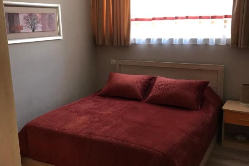 Un lit bien fait, avec une couverture rouge et des oreillers, un tableau encadré au mur et une fenêtre avec des rideaux.
