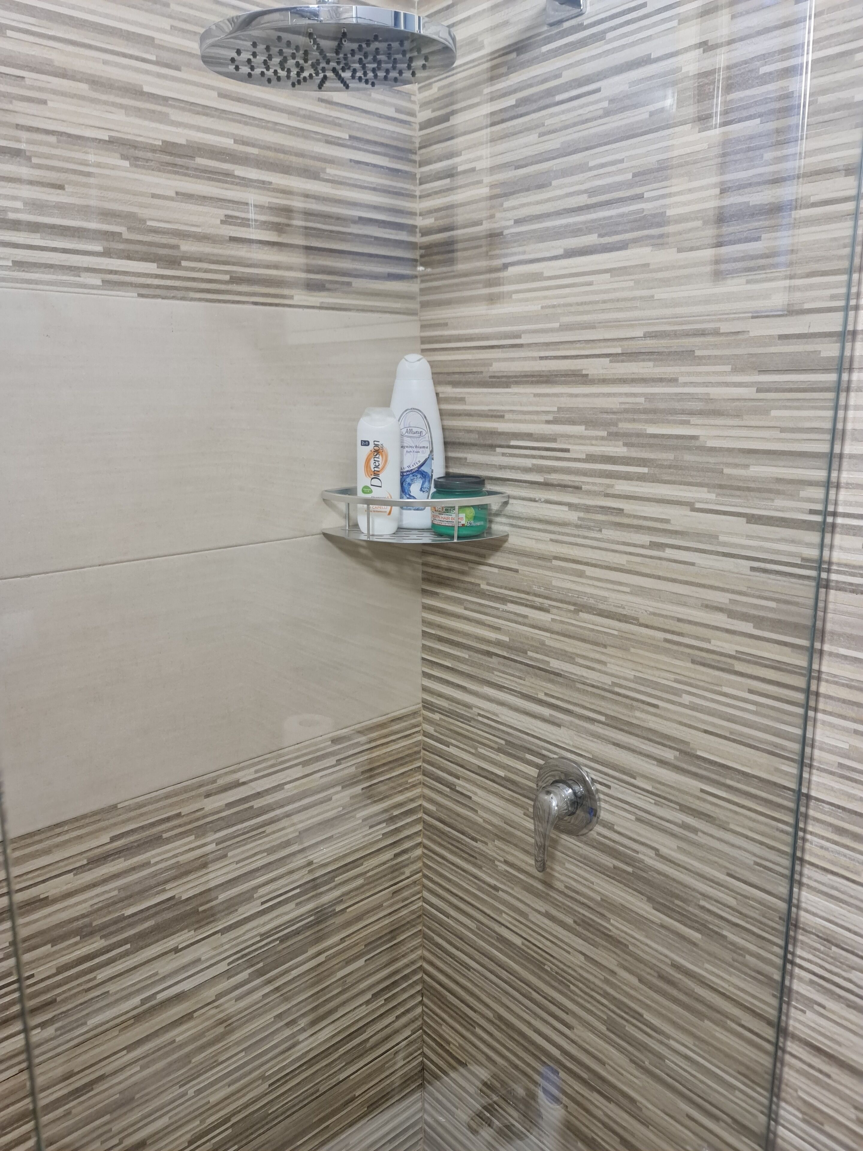 A shower area with a glass-enclosed showerhead, a shelf with toiletries, and a tiled wall.