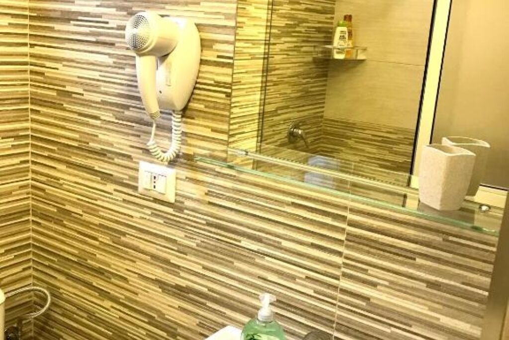 Un bagno con asciugacapelli montato a parete, cabina doccia in vetro e dispenser per sapone sul lavabo.