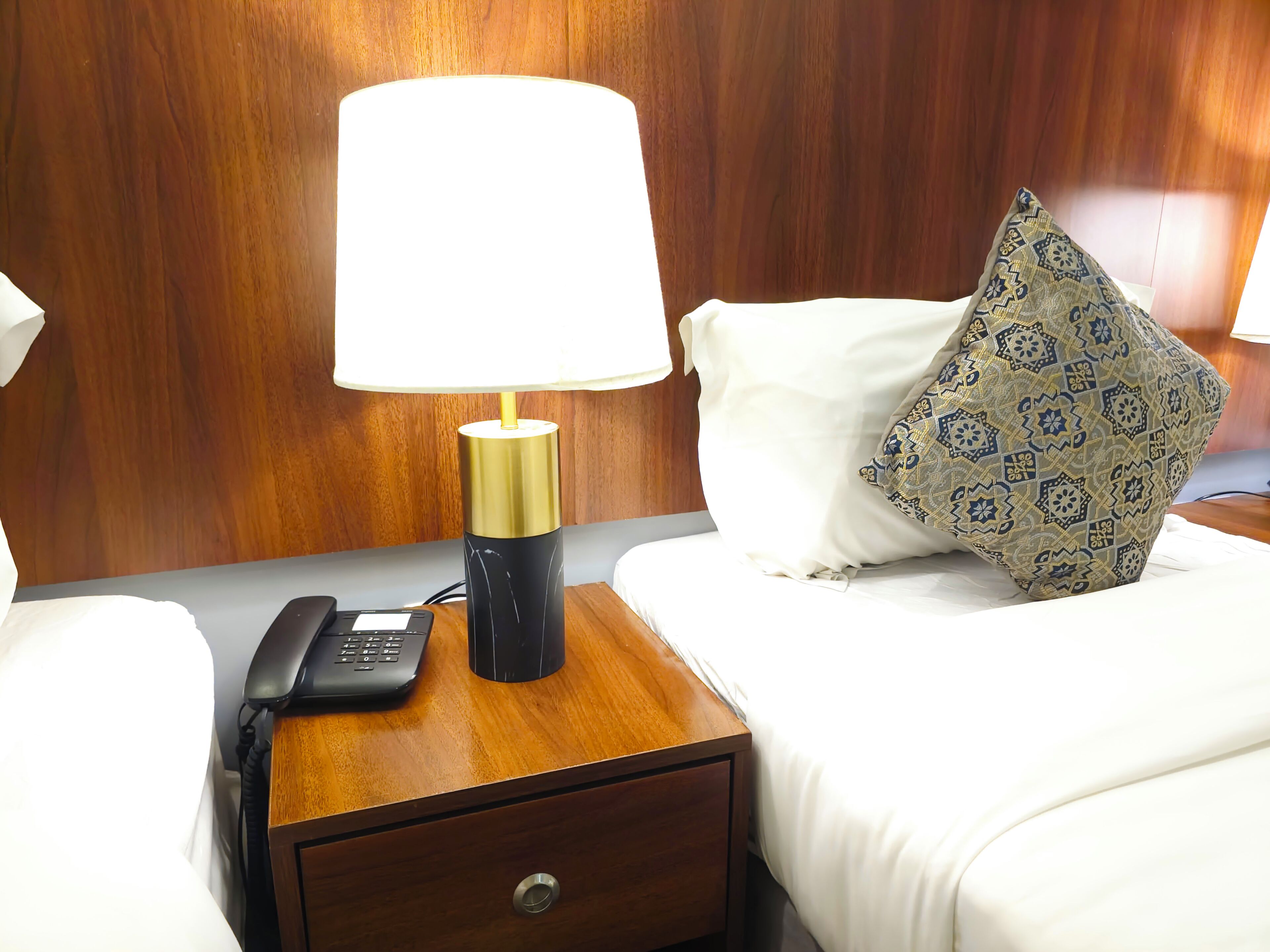 Habitación de hotel con cabecera de madera, una mesita de noche con lámpara, teléfono y almohadón decorativo.