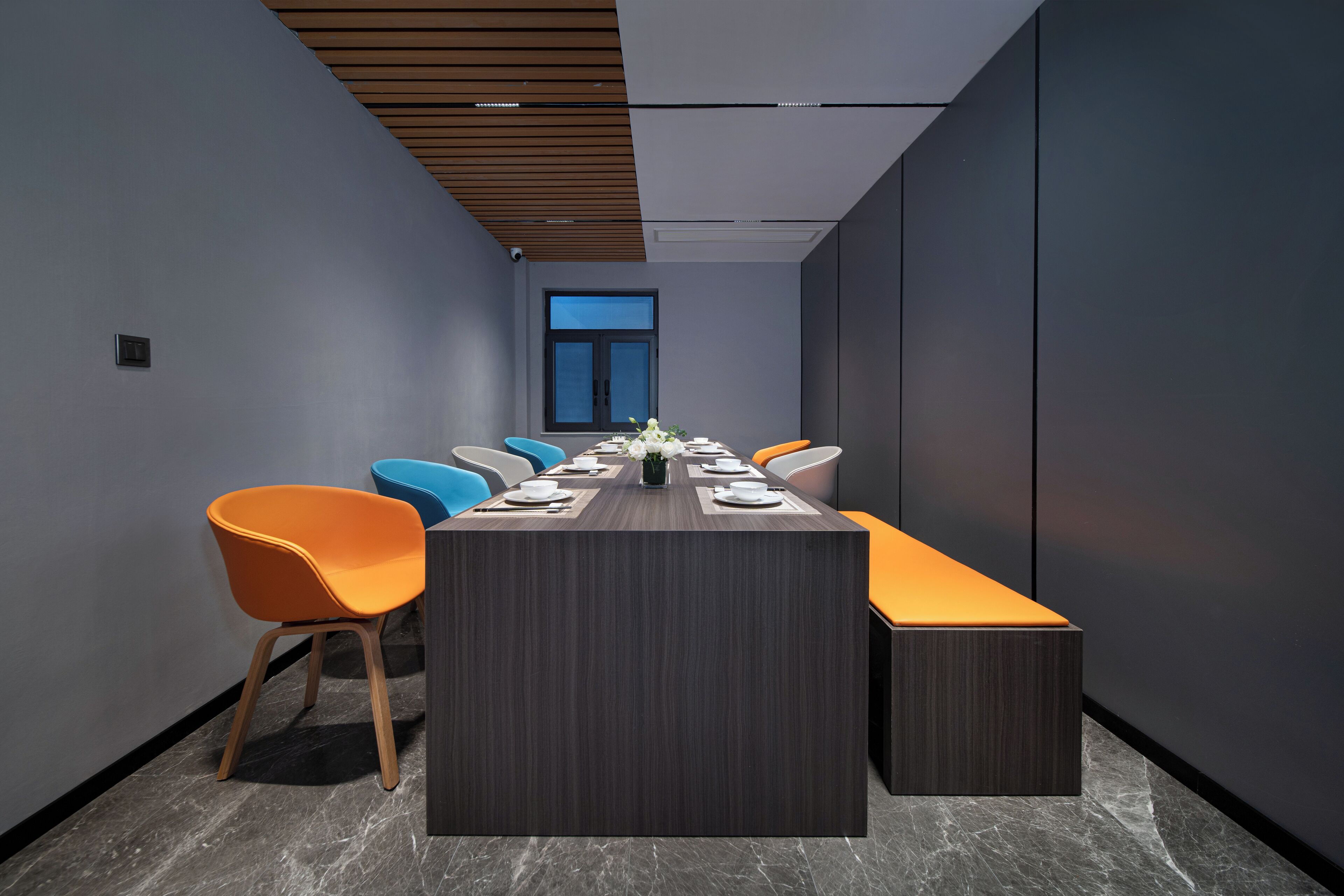 A modern conference room with a long table, orange and blue chairs, and a large window.