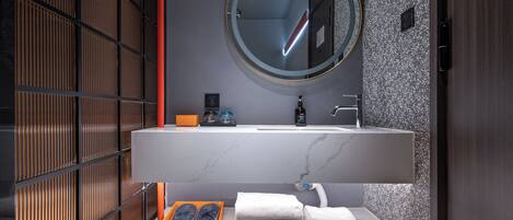 Een moderne badkamer met een houten vloer, een witte wastafel met een marmeren blad, een ronde spiegel en een handdoekhouder.