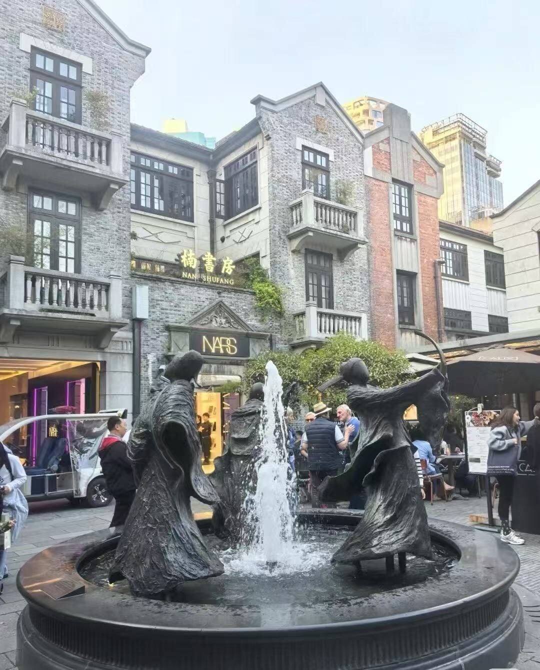 Una fuente pública con dos estatuas en el centro, rodeada por una plaza de piedra. Al fondo, se observan edificios de varios pisos con comercios.