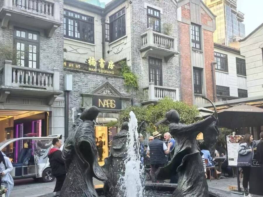 A public fountain with two statues in the center, surrounded by a stone plaza. In the background, there are multi-story buildings with commercial establishments.