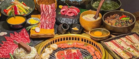 Variedad de alimentos, incluyendo sushi, sashimi y carnes a la parrilla, sobre una mesa con una parrilla central.