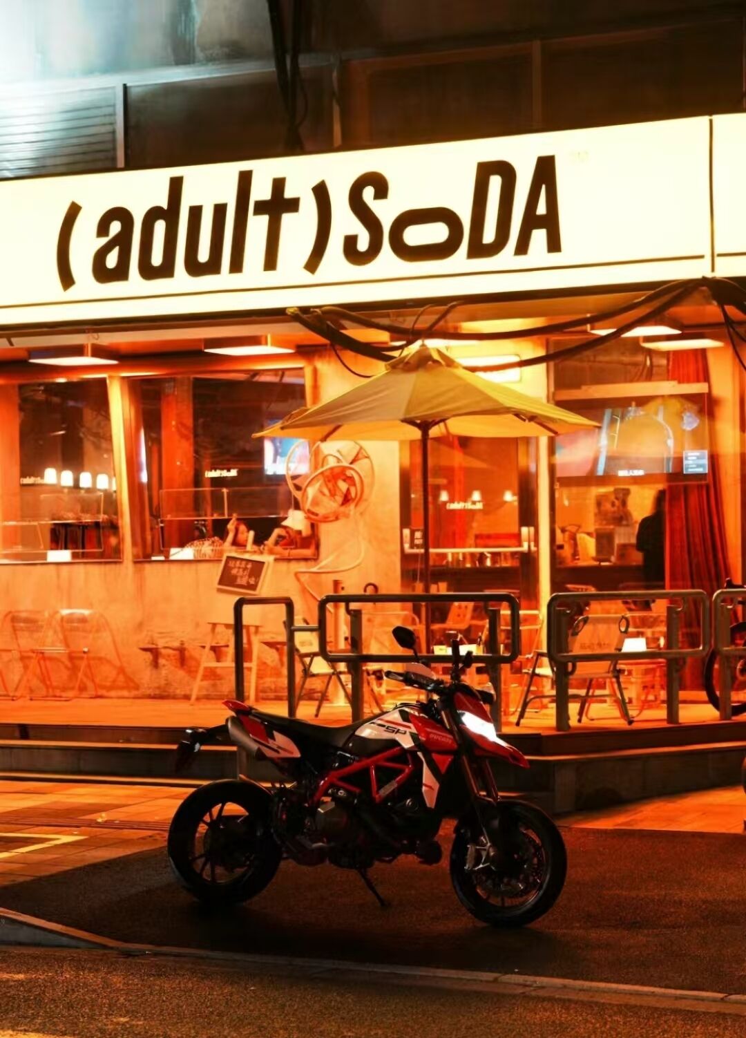 A motorcycle parked in front of a building with a sign that reads '(adult) Soda'.