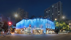 Un edificio moderno con fachada curva y paneles azules iluminados, situado en una intersección urbana con semáforos y vehículos a la vista.