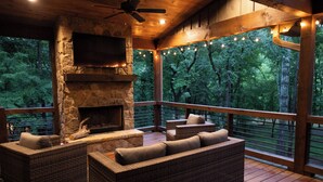 A covered outdoor seating area with wicker furniture, a stone fireplace, and a TV mounted on the wall. - Harmony River Retreat ~ Hot Tub ~ Fire Pit ~ Fish/Tube from the Backyard! (Ellijay)