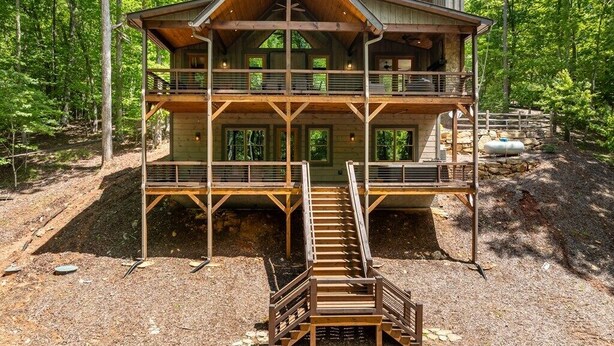A two-story log cabin with a balcony on the upper floor, surrounded by trees. - Harmony River Retreat ~ Hot Tub ~ Fire Pit ~ Fish/Tube from the Backyard! (Ellijay)
