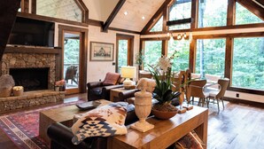 A spacious living room with a stone fireplace, wooden furniture, and large windows offering a view of the outdoors. - Harmony River Retreat ~ Hot Tub ~ Fire Pit ~ Fish/Tube from the Backyard! (Ellijay)