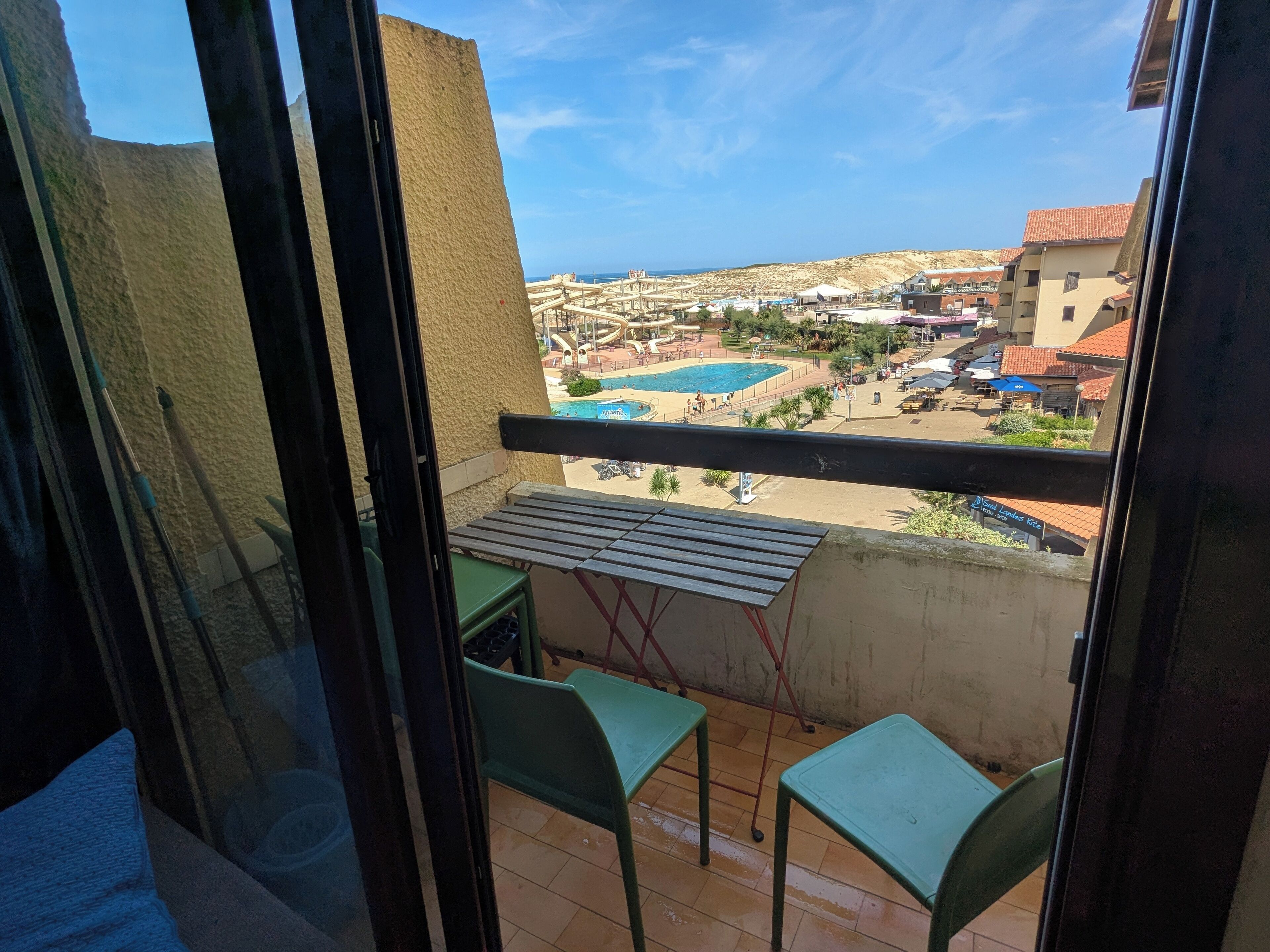 A balcony with two green chairs, a small table, and a view of a swimming pool and buildings in the distance.