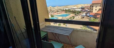 A balcony with two green chairs, a small table, and a view of a swimming pool and buildings in the distance.