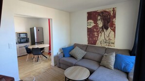 A living room with a grey sofa, blue cushions, a round coffee table, a kitchen area with a microwave and refrigerator, and a poster on the wall. - Beautiful T3 at the foot of the beaches, ocean view. (Seignosse)