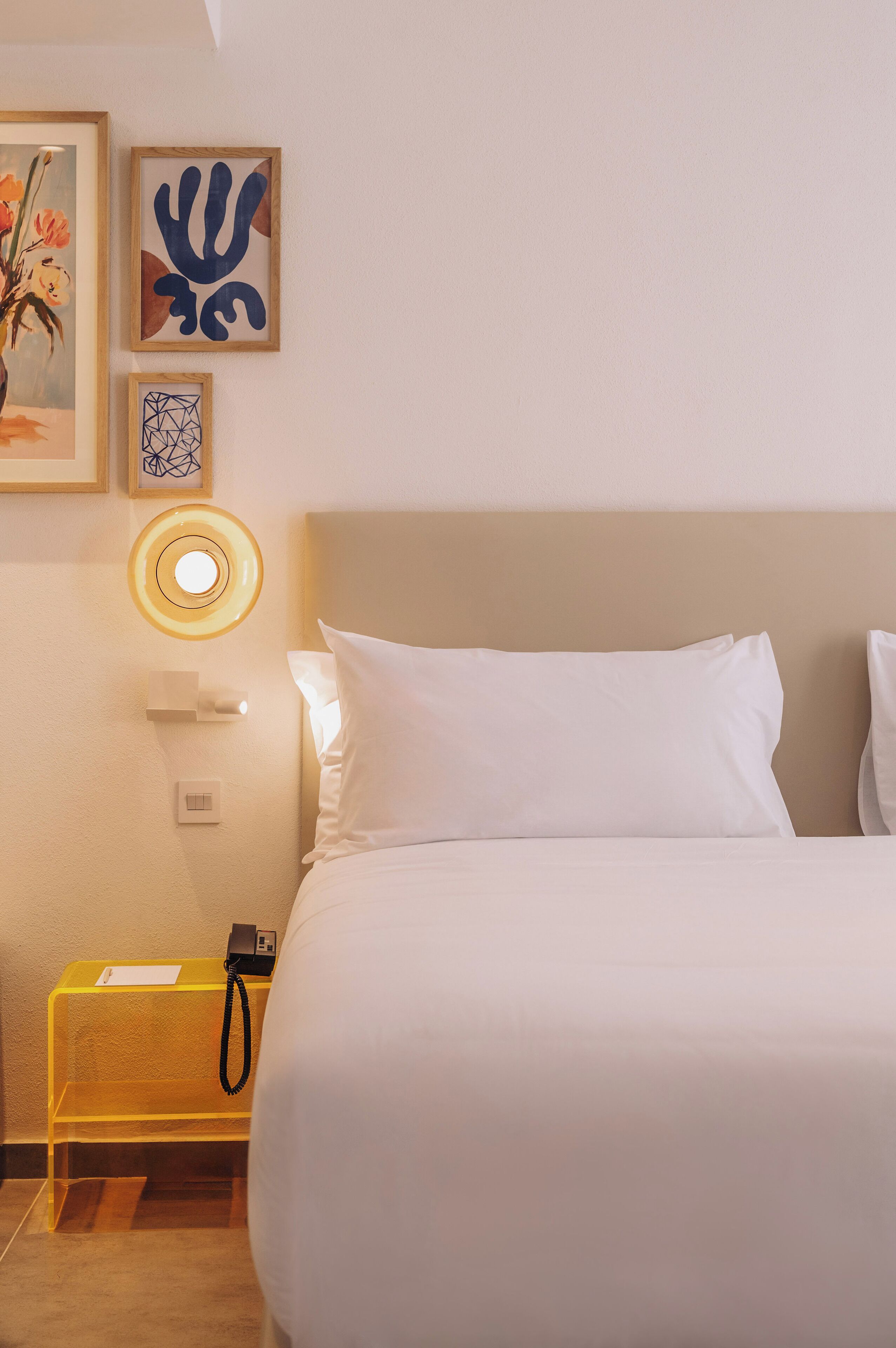 A hotel room with a bed, a bedside table with a lamp, and wall-mounted artwork.